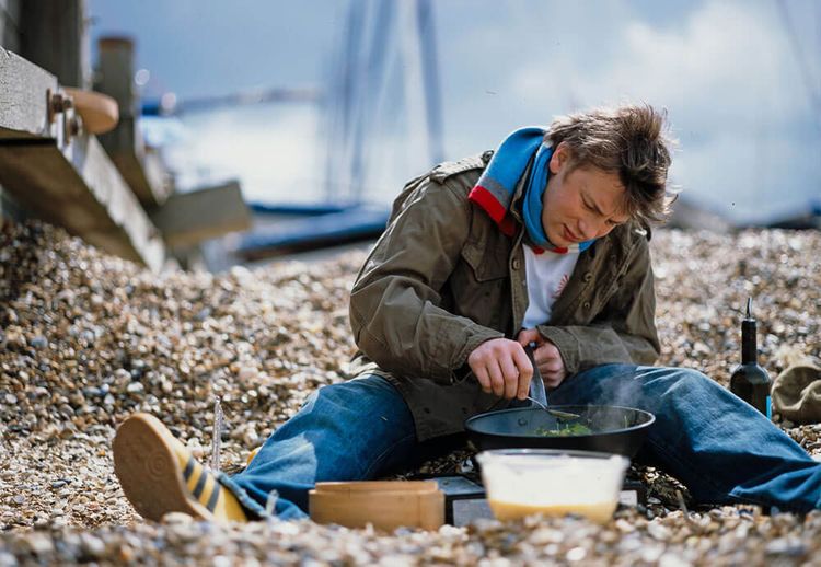 Jamie Oliver sitting cooking on a pebble beach