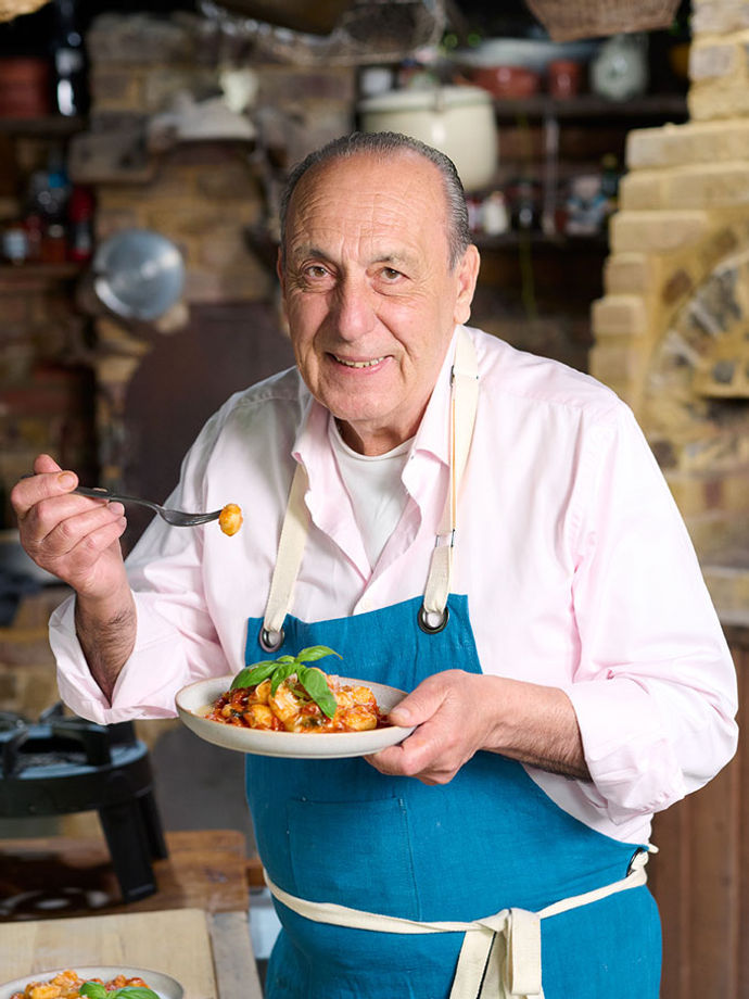 Gennaro Contaldo wearing a blue apron and holding a plate of fresh tomato gnocchi