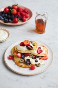 Fluffy pancakes with fruit and greek yoghurt, with a bowl of fruit and a glass of maple syrup above it