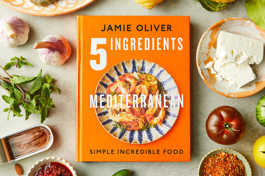 Jamie's cookbook 5 Ingredients Mediterranean - an orange hardback book surrounded by Mediterranean ingredients like garlic, feta, tomato and mint.