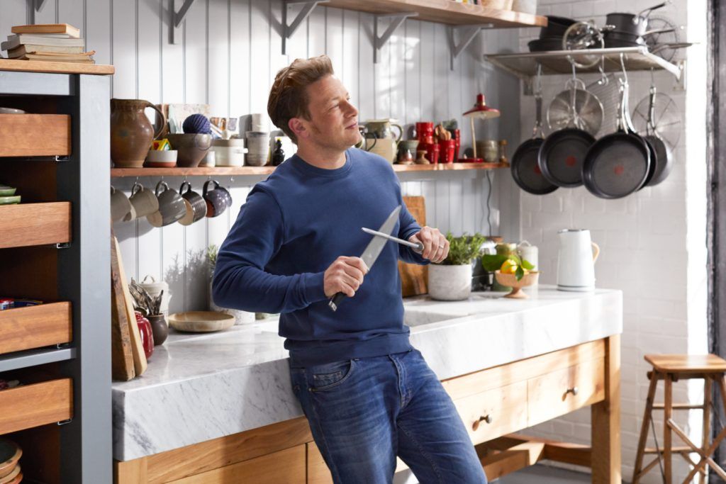 Jamie Oliver leaning against a table in a busy kitchen and looking to the side, while honing a knife using a steel. Picture from 'Jamie's Quick and Easy Food' TV show