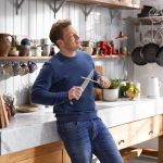 Jamie Oliver leaning against a table in a busy kitchen and looking to the side, while honing a knife using a steel. Picture from 'Jamie's Quick and Easy Food' TV show