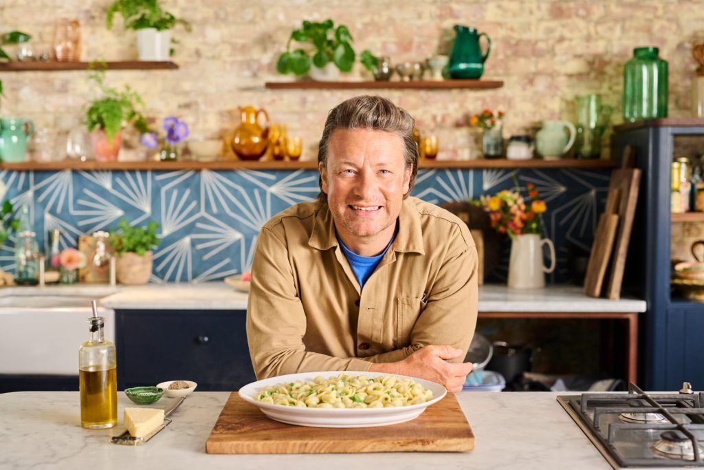 Jamie Oliver smiling at the camera with a large plate of % ingredient sweet pea orecchiette in front of him. A great midweek meal