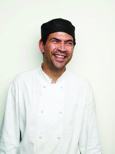 Almir Santos in his chef whites, smiling and stoof against a white wall